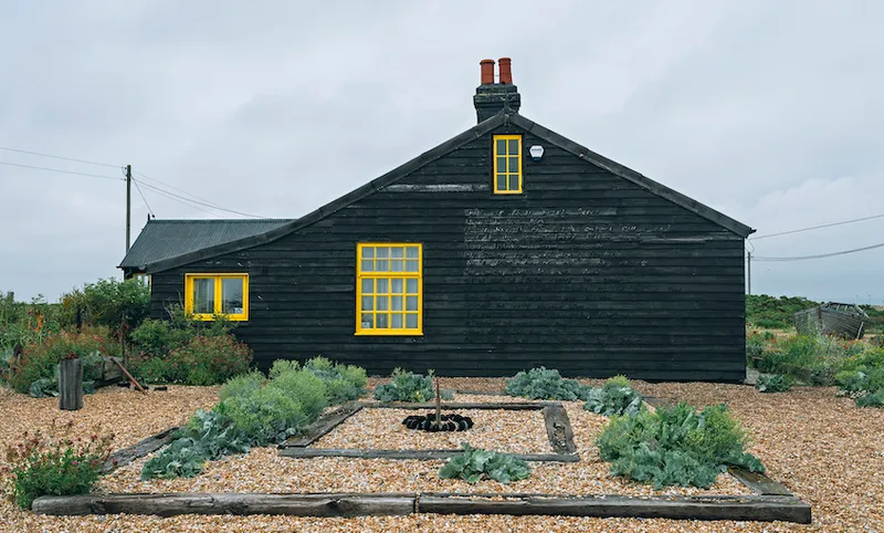 A photograph of a cottage, painted black with bright yellow window sills, in a striking garden of pebbles laid with sleepers and hardy plants.