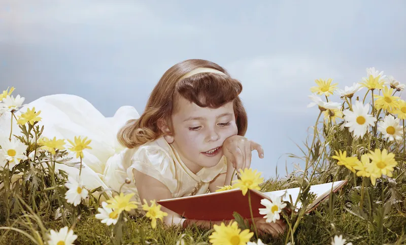 An old-timey photograph of a girl wearing a headband and lacey dress, lying in a dreamy meadow of daisies, reading a book. Bluebird sky.