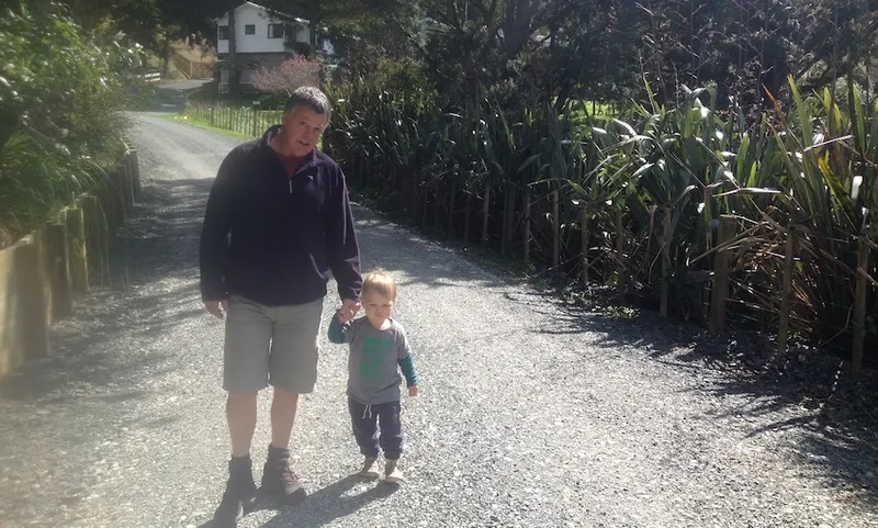 A middle-aged man holds a toddler by the hand, smiling, as they walk down a gravel driveway edged with harakeke. Beautiful day.
