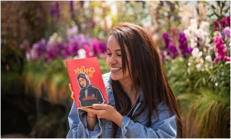 Young woman in garden holding novel The Pōrangi Boy, smiling