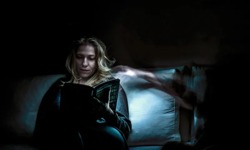 A dark room, a woman reads in bed, a ghostly arm reaching toward her