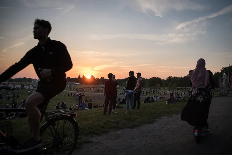 Sunset, a public park, we see groups of people, some walking, in the foreground a person on a bike