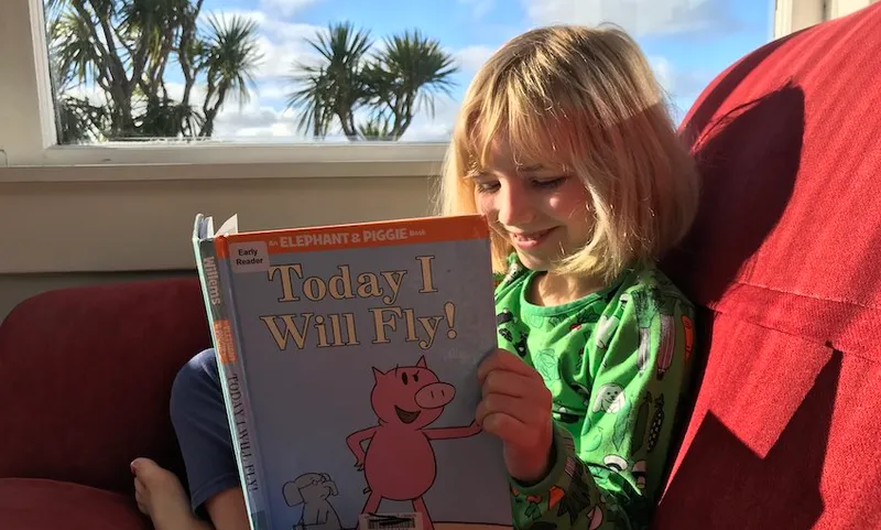 A blue-sky day, tī kōuka out the window, a blond, happy little girl sits on a bright red couch in the sun, reading.