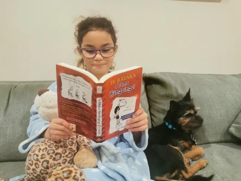 Young girl on a couch, a puppy beside her, she's wrapped up in a dressing gown and reading the te reo version of Diary of a Wimpy Kid.