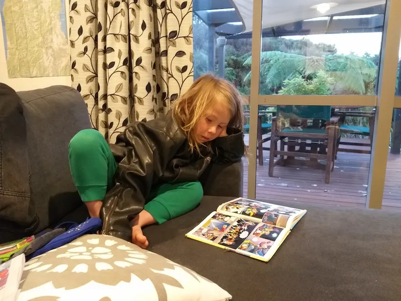 A child is curled in a corner on the floor, head propped up on elbow, riveted by the comic book on floor in front of him. Punga visible out the window.