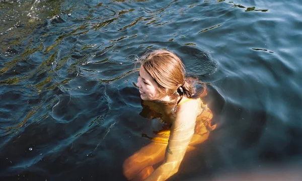 A young woman submerged in water up to her shoulders, dark water, outdoors, sun on her face, absolutely radiant
