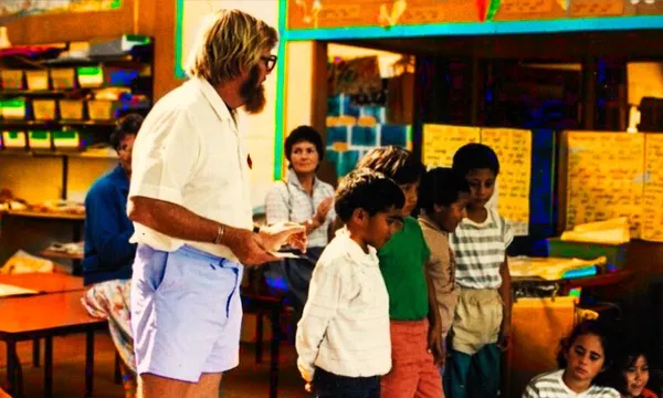 A 90s primary school classroom, male teacher with beard, short-sleeved shirt and walk shorts.