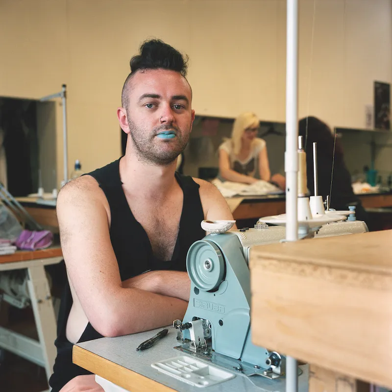 A young man wears a black singlet, blue lippy and a confident stare. He's seated at an industrial sewing machine, others (colleagues?) working in background.