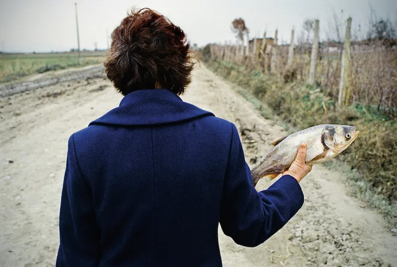 Odd photo of a woman holding a fish at shoulder height, pointing away from her body. She's walking on a gravel road, she has a coat on, we're watching her from behind.