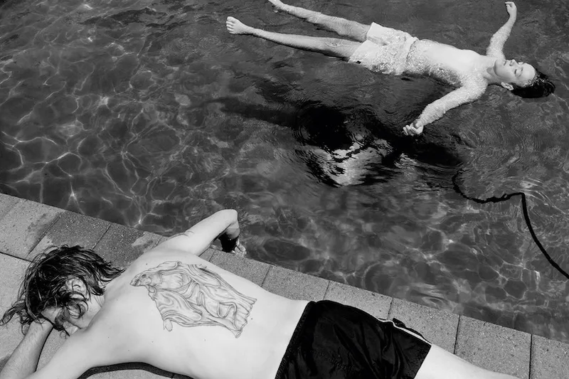 Black and white photograph: a child floats in a swimming pool, face-up, blissed out; a person lies face-down on the side of the pool, one hand dangling in the water.