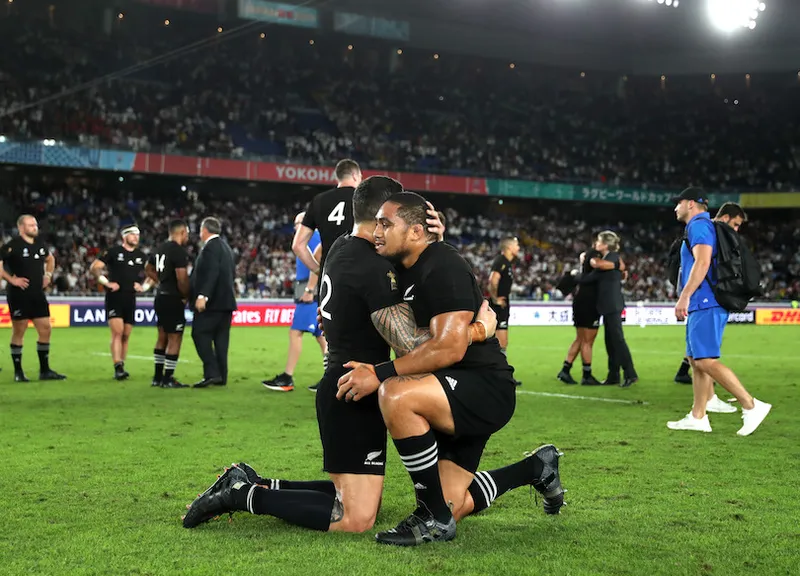 Sonny Bill Williams and Ofa Tu'ungafasi in the foreground, in All Blacks kit, embracing, on their knees; in the background, aftermath of a rugby match.