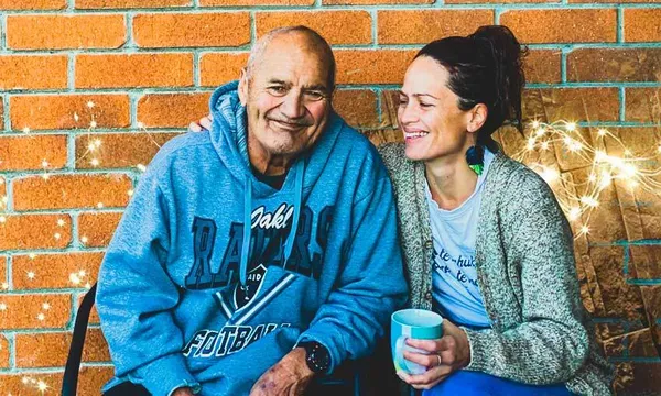 Two people sitting down in front of a brick wall. One is a woman she has her arm around an older man, her dad. They are smiling.
