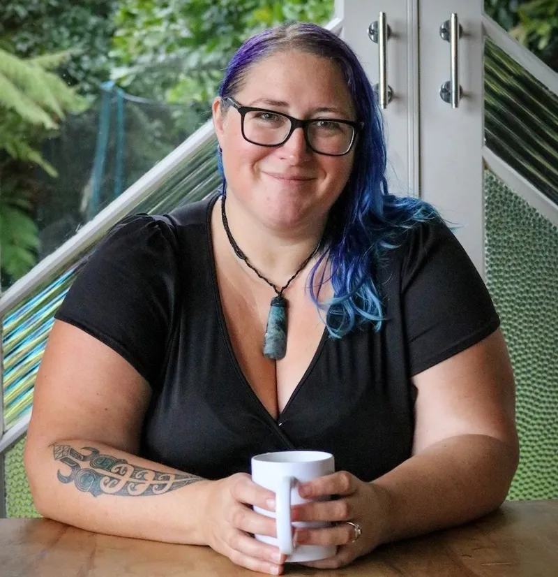 Smiling woman seated at a wooden table, hands wrapped round a cuppa, pounamu around her neck.