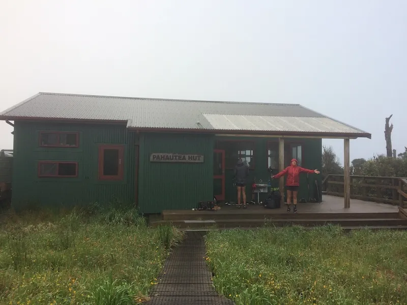 Exterior shot of a fancy-as DOC hut, a tramper standing triumphantly on the porch. Grey drizzly day.