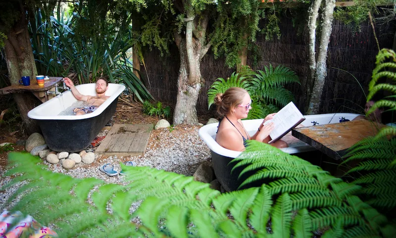 Idyllic setup: two baths in a garden of ferns and willow. In each one there's an adult relaxing; the woman in foreground is reading a large hardback.