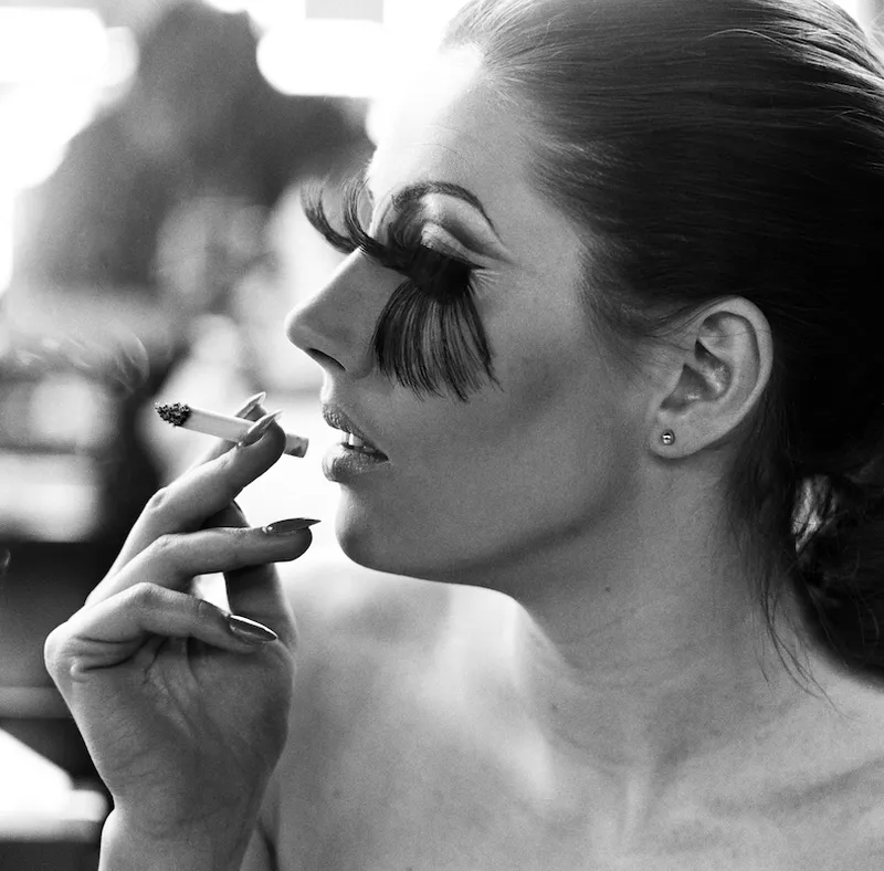 Black and white photo showing the profile of a young woman wearing ludicrously long false lashes – they reach her eyebrows and brush all the way down to the base of her nose. Smoking a cigarette, loving it.