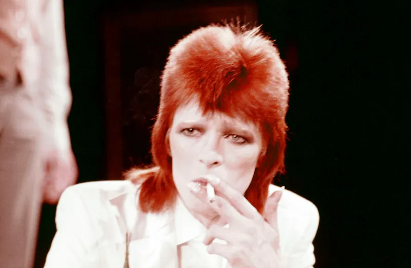 Pale, thin man, super glamorous, drags on a cigarette staring into middle distance. Bright orange mullet. White suit.