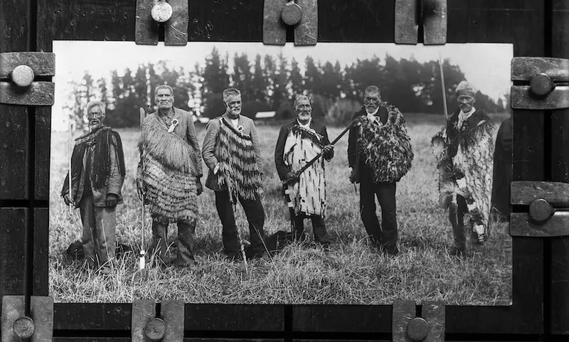 Black and white photograph of six Māori men standing in a row, facing the camera. All wearing various styles of kākahu over their suits.