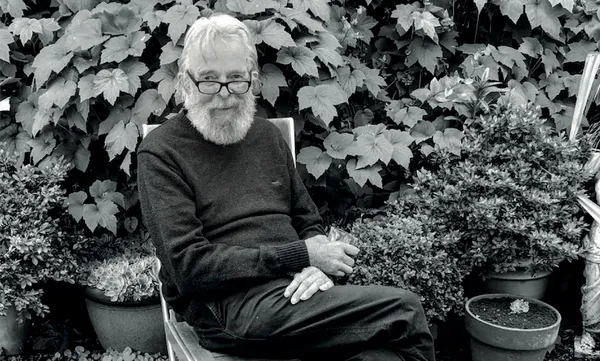 Black and white photograph: an older man, slight frame, switched-on look, seated in a garden. Legs crossed, he's holding a drink and looking to camera.