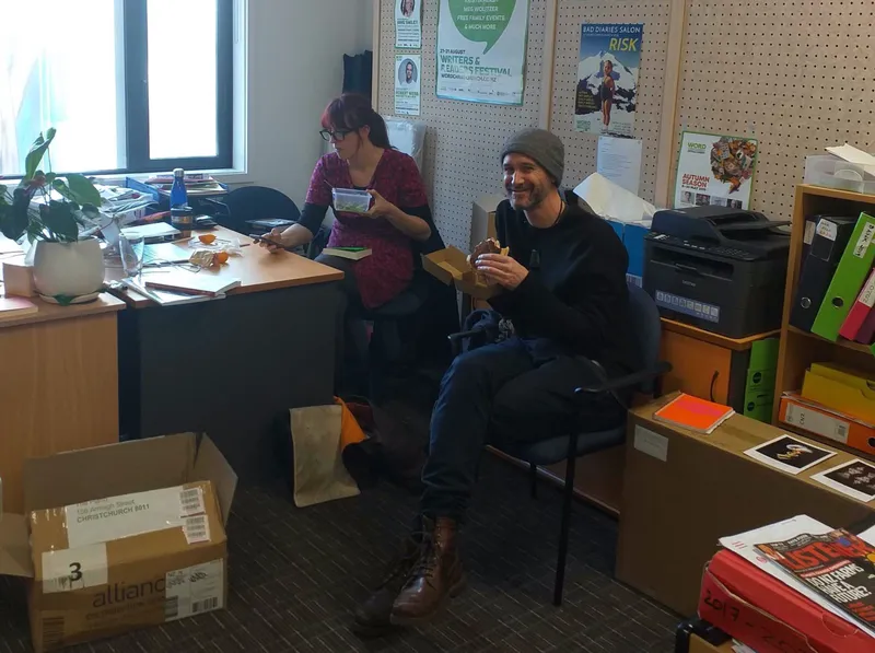 A thrown-together office room, boxes and bits and pieces everywhere, two people eating lunch.