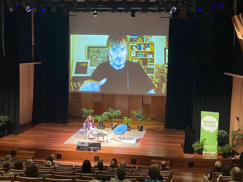 Photograph taken from far back in an auditorium, big screen up front on which a middle-aged man is talking; stage set up for host only.