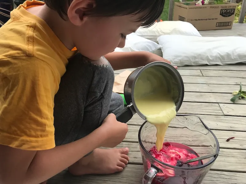 A boy on a wooden deck is tipping a saucepan of gloppy custard into a glass jug of radiant pink jelly.
