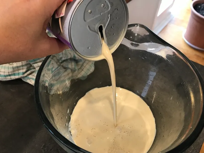 Pouring a can of evaporated milk into glass jug. Notably, the opening on the can is a long slit, rounded at top and bottom.