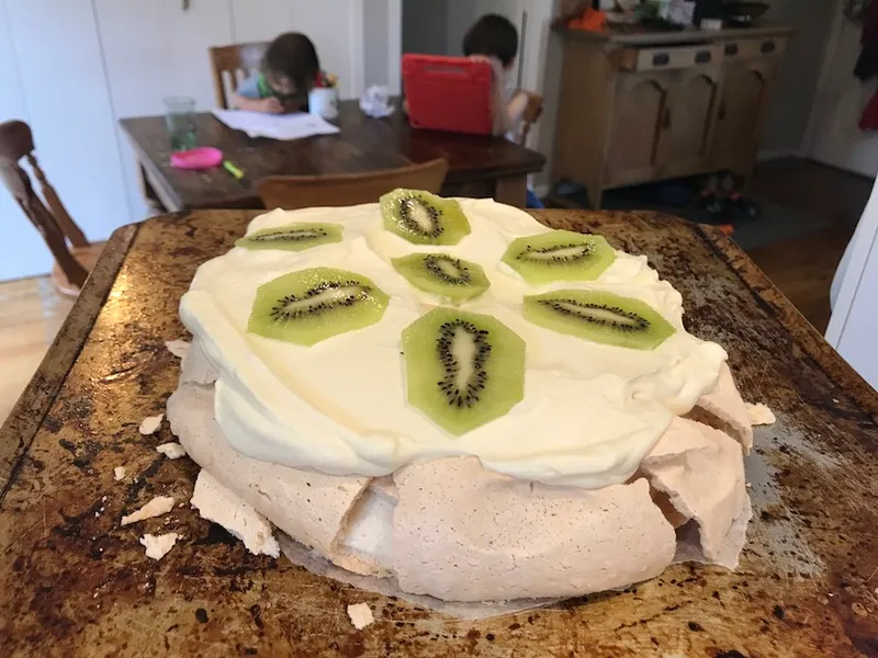 Background: kids drawing and on iPad at dining table. Foreground: a very flat pavlova, with cream and kiwifruit.