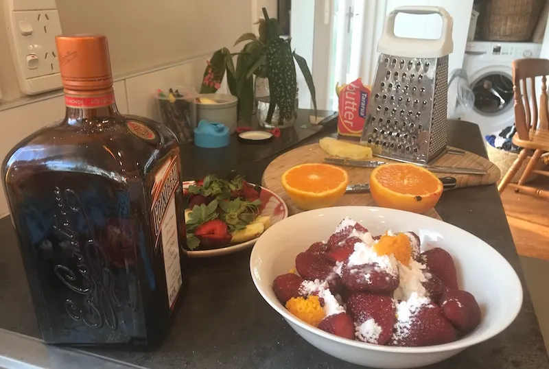 Kitchen bench strewn with stuff, including Cointreau, cut oranges, strawberry tops, spoons etc, a grater. Foreground: a bowl of strawberries covered in icing sugar and orange rind.