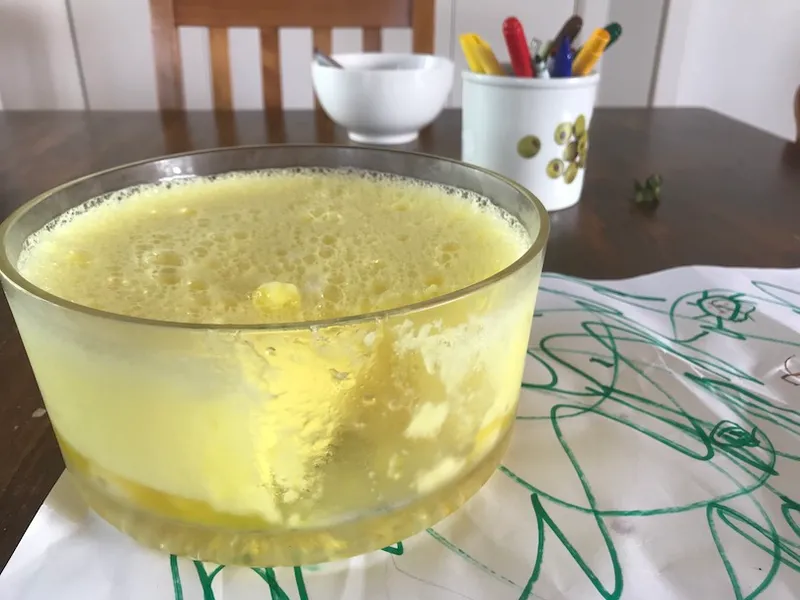 Side view of a luridly yellow dessert, fluffy on top with a layer of jelly at bottom. Typical little-kid detritus (felt pens, scribbled paper) all around.