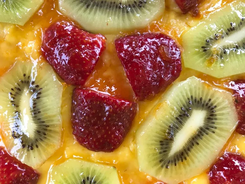 Aerial view of a section of a fruit flan. You see custard underneath, then a layer of sliced strawberries and kiwifruit, all covered in a slick apricot jam glaze.