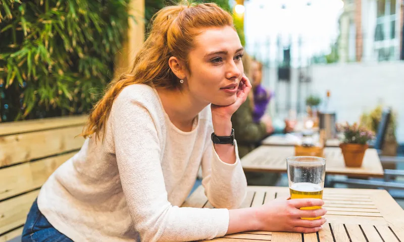 Woman sitting in pub garden, holding pint of beer, bored expression