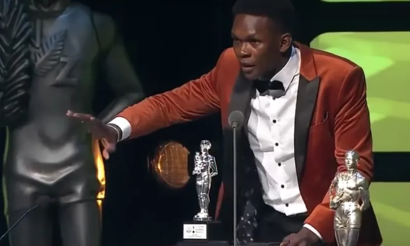 A man in a rust-colored tuxedo jacket stands at a podium, gesturing with one hand, while two trophies are placed in front of him. A silver statue is visible in the background.