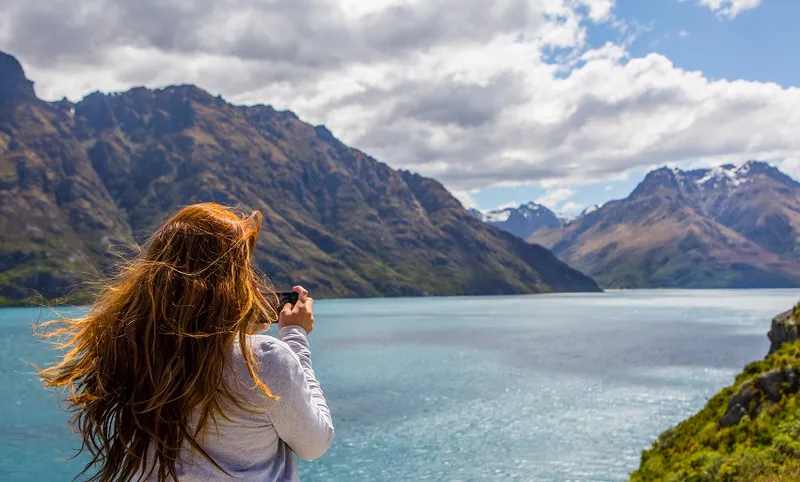 Caucasian woman photographing mountains and lake, Queenstown, Otago, New Zealand