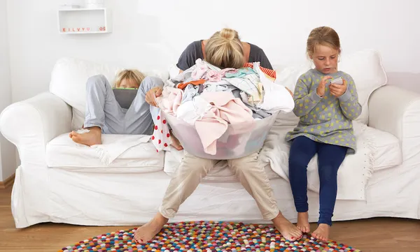 Exhausted mother with laundry basket on couch with children using digital tablet and cell phone