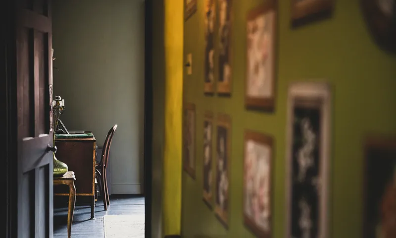 View along corridor with vintage pictures on green wall, open brown wooden door to study with antique desk and chair.