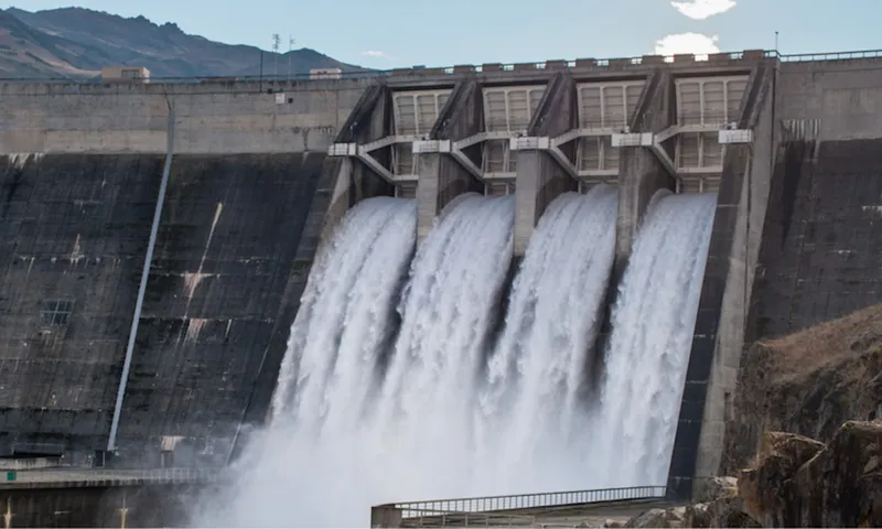 A large concrete dam releases water through four spillways, creating powerful streams. Mountains and a partly cloudy sky are visible in the background.