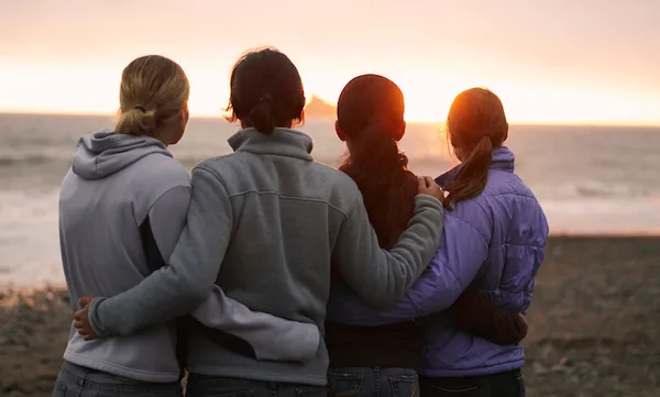Four young women, arms around each other, looking out to sea, rear view