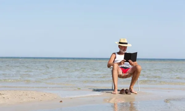 A man wearing shorts, T shirt and hat reads a book in a deckchair placed in the tide.