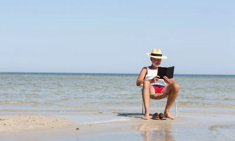A man wearing shorts, T shirt and hat reads a book in a deckchair placed in the tide.