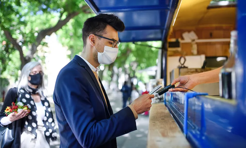 Portrait of young businessman with face mask buying coffee outdoors in city.