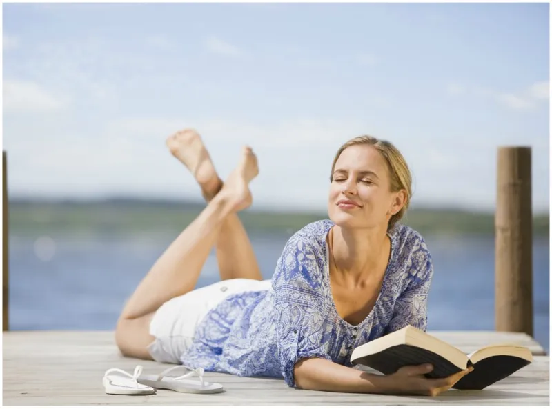 Woman lies tummy-down on a jetty, legs kicked up, propped on elbows with book.