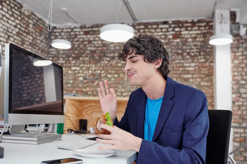 Young man sitting at desk, eating sandwich