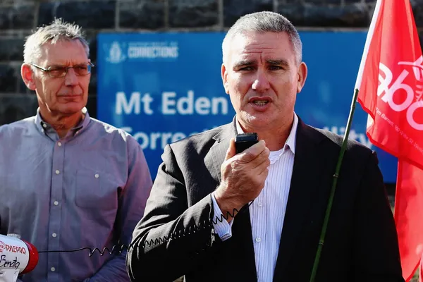 A man in a suit speaks into a microphone while holding a red flag, with another man in a buttoned shirt standing beside him. They are in front of a sign that reads "Mt Eden Corrections.