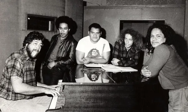 Five men gather around a grand piano in a recording studio. One plays the piano and smiles, while the others lean casually on the piano, looking at the camera. Sheet music is visible on the piano.
