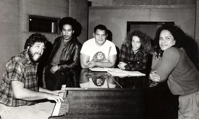 Five men gather around a grand piano in a recording studio. One plays the piano and smiles, while the others lean casually on the piano, looking at the camera. Sheet music is visible on the piano.