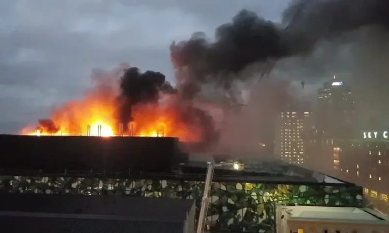 A large fire burns on the roof of a building at night, with thick black smoke rising into the sky; city skyscrapers with lights are visible in the background.