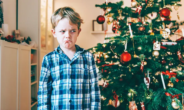 Unhappy boy stands before chistmas tree and makes a face