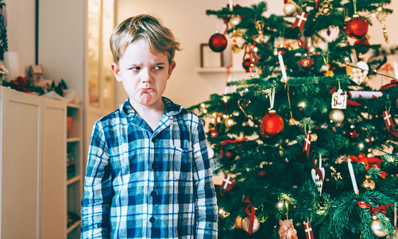 Unhappy boy stands before chistmas tree and makes a face