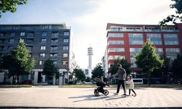Father walking with baby stroller looking at daughter riding push scooter in city